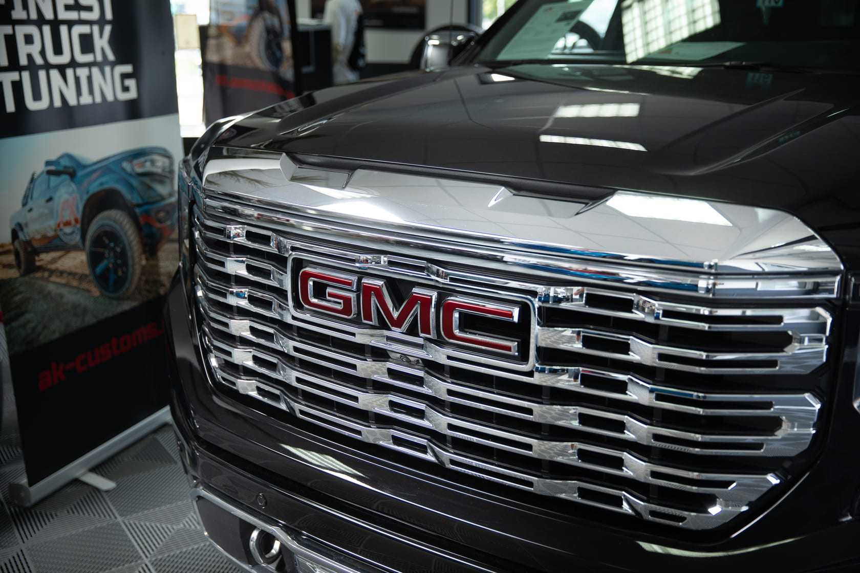 Das Bild zeigt eine Nahaufnahme des eindrucksvoll verchromten Kühlergrills des 2025 GMC Sierra 1500 Denali, der mit seinem markanten roten GMC-Logo in der Mitte sofort ins Auge fällt. Die horizontalen Chromlamellen spiegeln das Licht der Showroom-Decke und unterstreichen den luxuriösen Charakter des Fahrzeugs. Links im Hintergrund ist ein Banner von AK Customs zu sehen, das den Bezug zum professionellen Truck-Tuning herstellt.